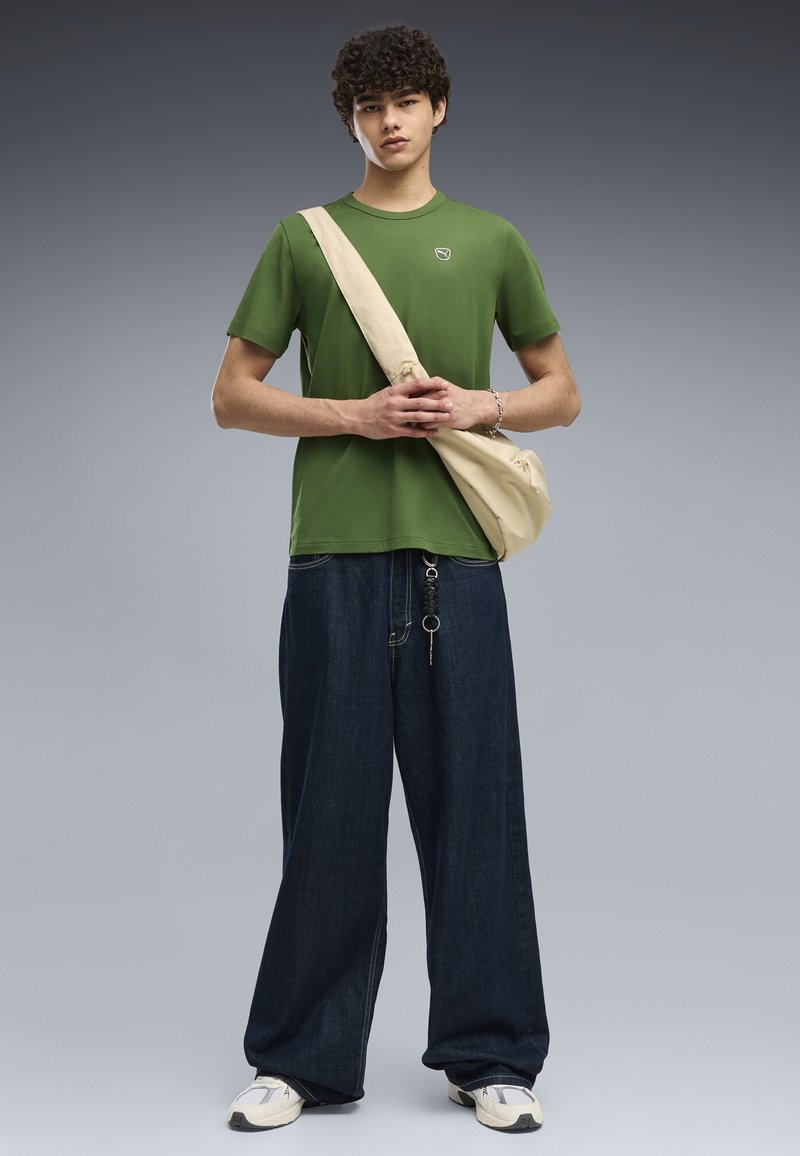 Jeune homme aux cheveux bouclés portant un T-shirt vert, un jean large foncé, des baskets blanches et un sac bandoulière beige, debout devant un fond uni.