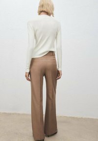 Personne avec les cheveux blonds courts portant un haut à manches longues blanc et un pantalon marron large taille haute, se tenant devant un mur uni.