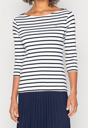 Kvinde iført en hvid og marineblå stribet langærmet skjorte med et lille logo på brystet og et navy plisseret skørt.