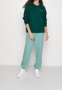 Sudadera con capucha de color verde oscuro con bolsillo frontal y cordón ajustable. Pantalones deportivos azul claro con puños elásticos. Se lleva con zapatillas blancas.