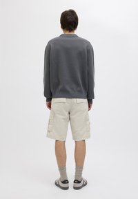 Grauer strukturierter Sweatshirt, beige Cargo-Shorts mit seitlichen Taschen, hellgraue Socken und Sandalen. Modell von hinten vor einem weißen Hintergrund betrachtet.