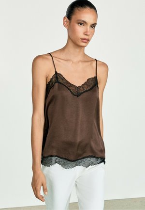 Donna che indossa una canottiera in raso marrone con bordatura in pizzo nero e spalline sottili, abbinata a pantaloni bianchi, in piedi davanti a uno sfondo semplice.