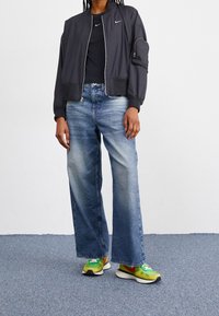 Zwarte bomberjack met elastische manchetten, lichtblauwe wijde jeans en groene, gele, rode Nike sneakers. De outfit heeft een zwart topje eronder.