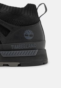 Talons de chaussures Timberland noires avec col de cheville en tricot, languette de tirage, semelle texturée et logo Timberland gris avec marquage.