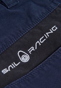 Tissu bleu marine et noir avec des textures contrastées. Présente un logo brodé blanc "Sail Racing" et un design en spirale. Vue en gros plan.
