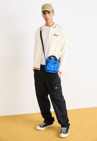 Saco de ombro azul, cardigan branco com detalhes vermelhos, t-shirt branca simples, calças cargo pretas e ténis pretos. Fundo amarelo.