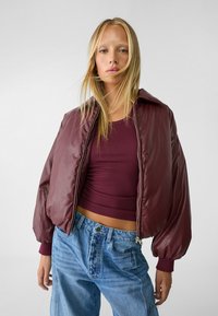 Burgunderfarbene Bomberjacke aus glänzendem Nylon mit Reißverschluss, gerippten Bündchen und voluminösen Ärmeln, kombiniert mit einem figurbetonten burgunderfarbenen Oberteil und blauen Jeans.