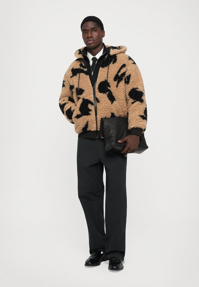 Man in een beige en zwart gestreepte sherpa jas, zwarte broek, zwarte schoenen, met een zwart leren clutch voor een witte achtergrond.