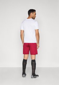 Weißes sportliches Shirt mit kurzen Ärmeln, kombiniert mit roten Shorts, schwarzen knielangen Socken und schwarzen Fußballschuhen. Einfaches Design, sportliche Passform.