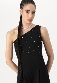 Black one-shoulder dress made of smooth fabric, adorned with white pearl embellishments, featuring a fitted bodice and pleated skirt.