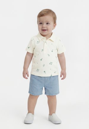 Dz toddler stoi, ubrany w kremową koszulkę polo z zielonymi wzorami, jasno niebieskie spodenki i białe buty wsuwane, lekko się uśmiecha.