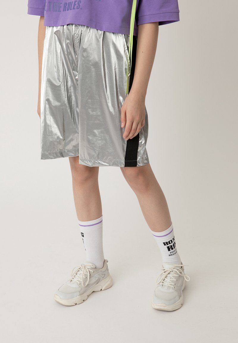Pantaloni corti argentati e lucidi con una vestibilità ampia e un accento nero di lato, abbinati a calze bianche con strisce viola e sneaker grigio chiaro.