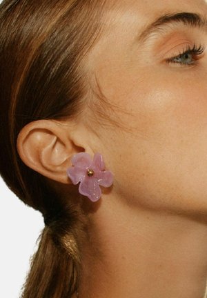 Stranski profil ženske, ki prikazuje bledo vijolično prozorno uhan v obliki cvetlice z zlatim središčem na ušesu, svetlo rjavi lasje, zavezani nazaj.