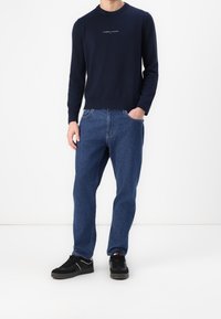 Granatowy sweter na drutach z prążkowanymi mankietami i dołem, w zestawie z luźnymi niebieskimi dżinsami. Czarnie trampki dopełniają strój.
