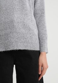 Person wearing a light gray knitted sweater and black pants, with their right hand relaxed beside their hip.