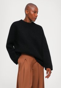 Nainen, joka on pukeutunut mustaan ylisuurta neuleeseen ja korkeavyötäröisiin, laskostettuihin ruskeisiin housuihin, seisoo yksi käsi taskussa tasaisella taustalla.