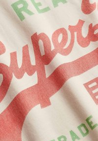 Gros plan d'un tissu avec "Superdry" en grande écriture rouge et texte vert partiel sur fond crème.
