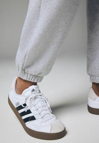 Weiße Sneaker mit schwarzen Streifen, Wildleder-Zehenkappe und brauner Gummisohle. Graue, tapered Sweatpants mit elastischen Bündchen.
