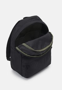 Sac à dos en tissu noir avec un rabat ouvert révélant un intérieur bordé de vert et plusieurs compartiments pour un rangement organisé. Matériau texturé.