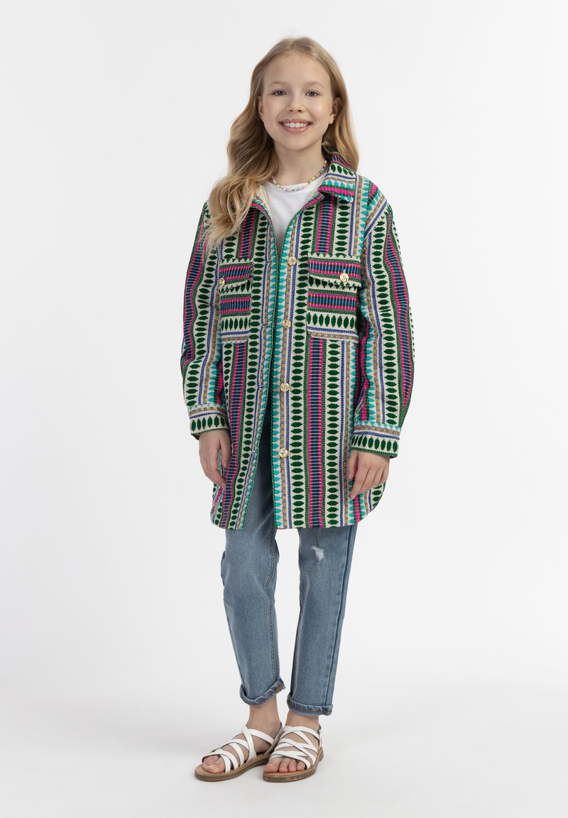 Värikäs pitkä takki vihreästä, pinkistä ja sinisestä kuviollisesta kankaasta, jossa on kaksi etutaskua, yhdistettynä vaaleansininisiin farkkuihin ja valkoisiin sandaaleihin.