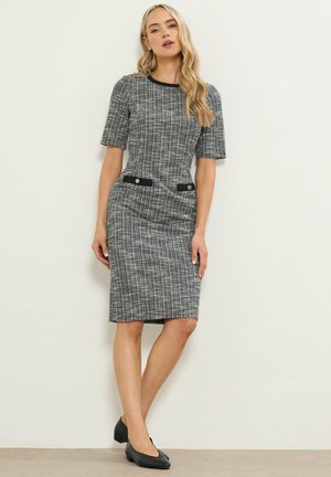 Grå tweedkjole med korte ærmer, tætsiddende silhuet, sort kant og to sorte knapdetaljer i taljen. Båret med sorte flade sko.