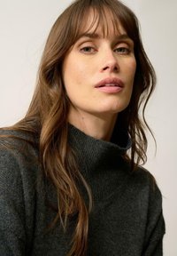 Szary sweter z dzianiny o wysokim kołnierzu, z miękką strukturą i warstwowym wyglądem, w połączeniu z długimi, falującymi brązowymi włosami na neutralnym tle.