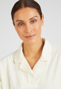 Camisa de corduroy blanco con cuello extendido, que presenta dos botones y detalles de costura visibles. Textura suave y apariencia ligera.