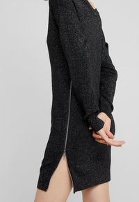 Abito in maglia nera e luccicante con maniche lunghe, scollatura a girocollo e zip laterale. Il design presenta uno spacco alto sul lato sinistro.