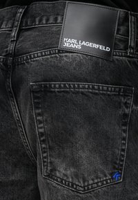 Jeans de ganga pretos com um acabamento texturizado. Apresenta um patch de couro com a marca e uma pequena costura azul na parte de trás do bolso.