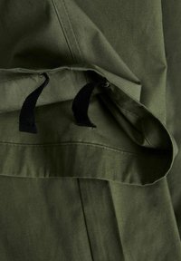 Tissu vert olive avec deux cordons courts noirs suspendus à une couture ouverte ou au bord d'une poche.