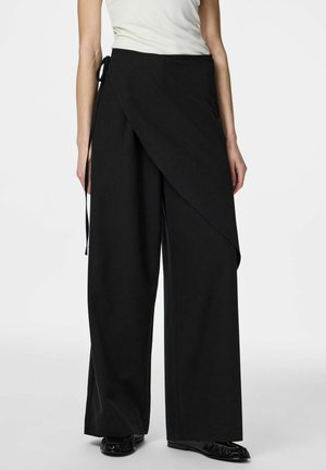 Femme portant un pantalon noir à jambes larges avec un détail de superposition noué à la taille, associé à un haut blanc sans manches et des chaussures noires.