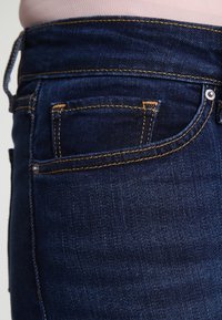 Los vaqueros de denim azul oscuro presentan una vista en primer plano del bolsillo trasero con costuras amarillas y un acento de remache plateado.