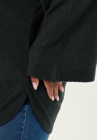 Schwarzer gerippter Pullover mit weiten Ärmeln. Seitlicher Schlitz am Saum. Nahaufnahme von Fingern, die in der Tasche ruhen. Strukturierter Stoff, weiche Optik.