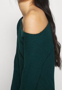 Pullover lavorato a maglia verde scuro con design off-the-shoulder, caratterizzato da una texture a coste e maniche ampie.