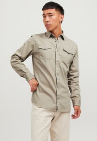 Camisa de manga larga color beige claro con dos bolsillos en el pecho, cierre frontal con botones y broches a presión. Textura de tela suave con un diseño ajustado.