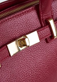 Close-up van een leerachtige rode handtas met textuur, voorzien van een goudkleurige draaiklink en een zichtbare rits met gouden tanden.