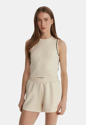 Donna con capelli castani lunghi, indossa un top corto beige senza maniche e pantaloncini coordinati a vita alta, in piedi davanti a uno sfondo semplice.