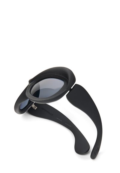 Black sunglasses with a rounded design, featuring dark lenses and textured frames. Thin, flexible arms extend from the main body.