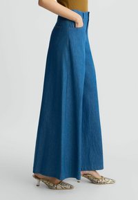 Hochtaillierte, weit geschnittene blaue Denim-Hosen mit zwei Vordertaschen. Kombiniert mit gemusterten Schuhen mit spitzer Zehe und kleinem Absatz.