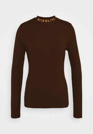 Brun långärmad topp med hög hals, med orange texten "STIEGLITZ" vid kragen. Slätt, figurnära tyg med minimal detaljering.