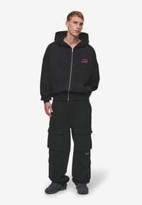 Čierna mikina na zips s ružovým logom, v kombinácii s čiernymi cargo nohavicami s viacerými vreckami. Model má na sebe tmavé športové topánky.