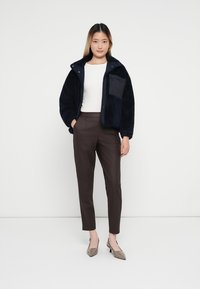 Giacca di pile blu navy con colletto alto e taschino sul petto, abbinata a pantaloni marroni su misura e tacchi marrone chiaro, su uno sfondo neutro.