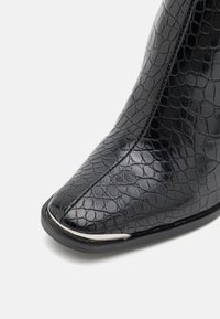 Botte noire à la cheville en simili cuir crocodile brillant, avec un bout pointu et un détail argenté le long de la semelle. Texture lisse.