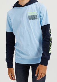 Pojke som bär en ljusblå kortärmad skjorta över en mörkblå långärmad tröja med neontexten "SLIDING RAMPS GRINDING RAILS" och "FLIP THE BOARD."