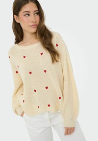 Jeune femme portant un pull en tricot crème avec des cœurs rouges, un pantalon blanc, les cheveux longs et ondulés, regardant sur le côté sur un fond uni.