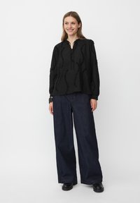 Blusa nera con ruche texturizzate, maniche lunghe e scollatura a V, abbinata a jeans in denim scuro a gamba larga e scarpe nere.