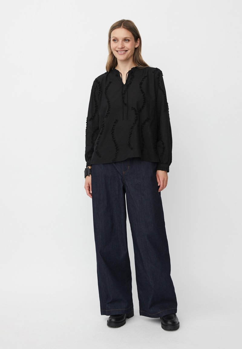 Blusa nera con ruche texturizzate, maniche lunghe e scollatura a V, abbinata a jeans in denim scuro a gamba larga e scarpe nere.