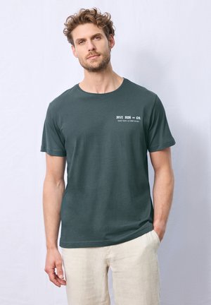 Dark green cotton t-shirt with short sleeves, featuring a printed logo on the left chest. Paired with light-colored linen shorts.
