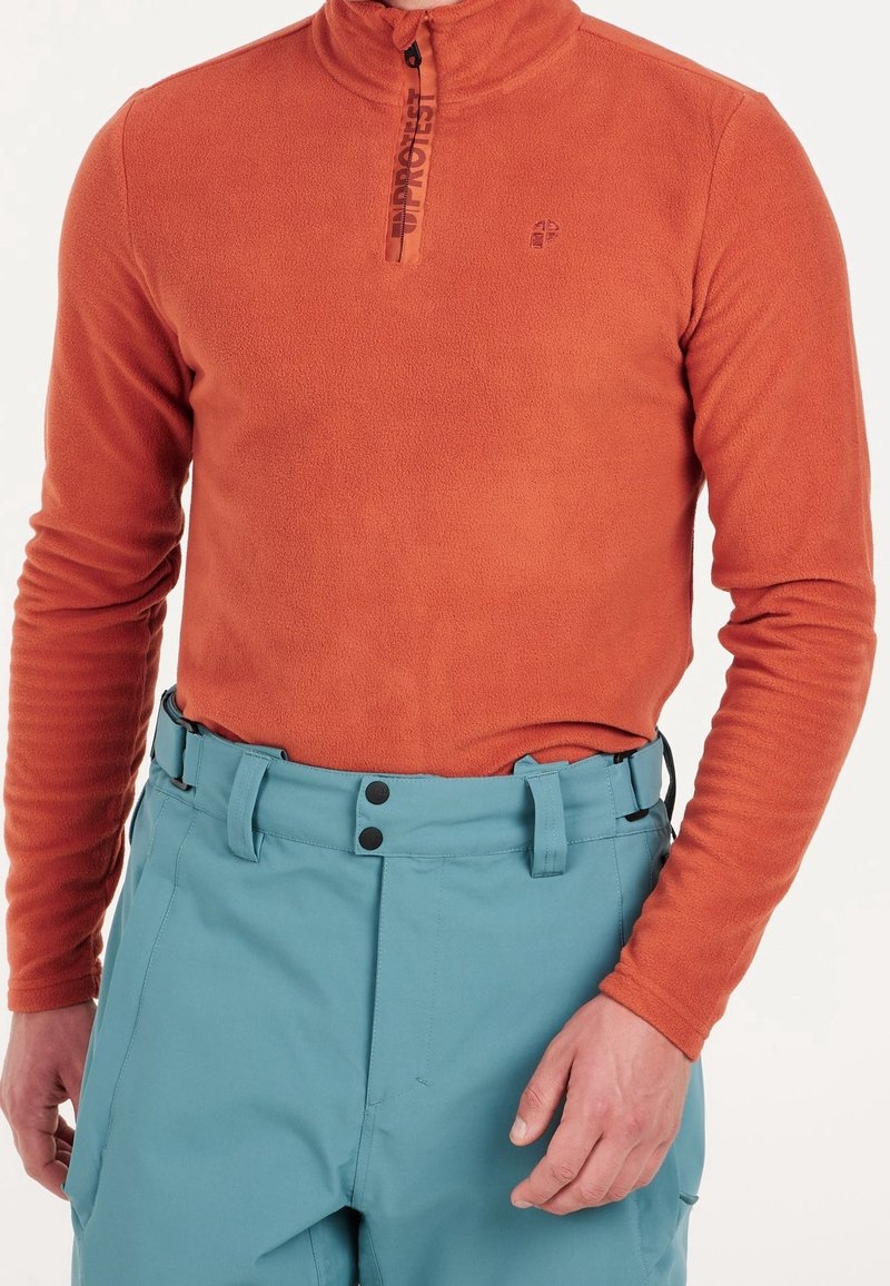 Homme portant un pull polaire orange à manches longues avec une fermeture éclair au quart et un pantalon taille haute bleu clair avec boutons.