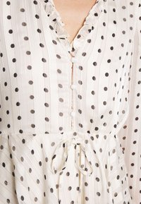 Blusa blanca con lunares negros, con un escote fruncido, cierres de botones y un detalle de lazo en la cintura. Tela suave y fluida.